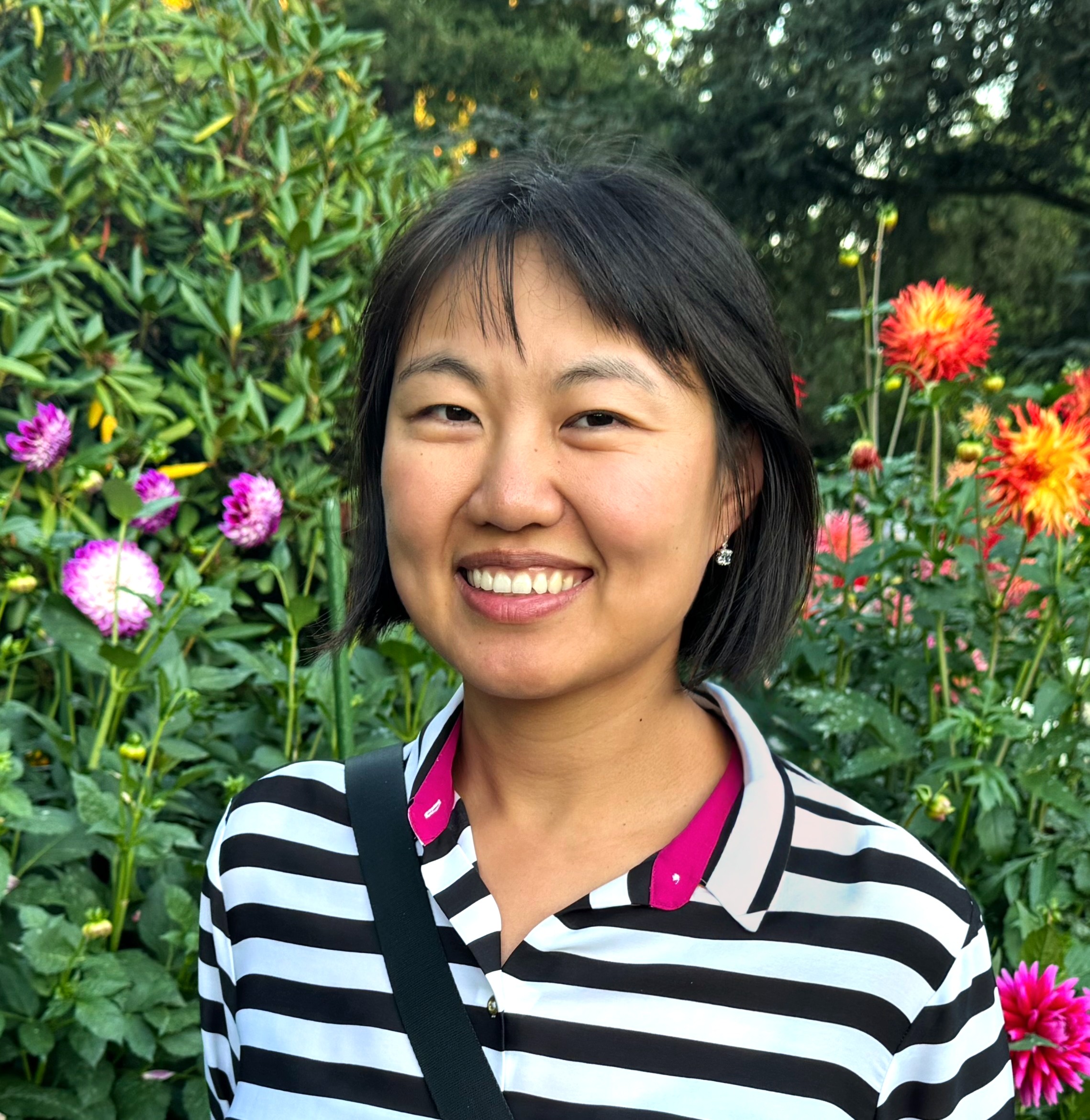 portrait photo of a smiling east asian woman with chin-length black hair, wearing a black and white striped shirt, in front of a background of trees and dahlias