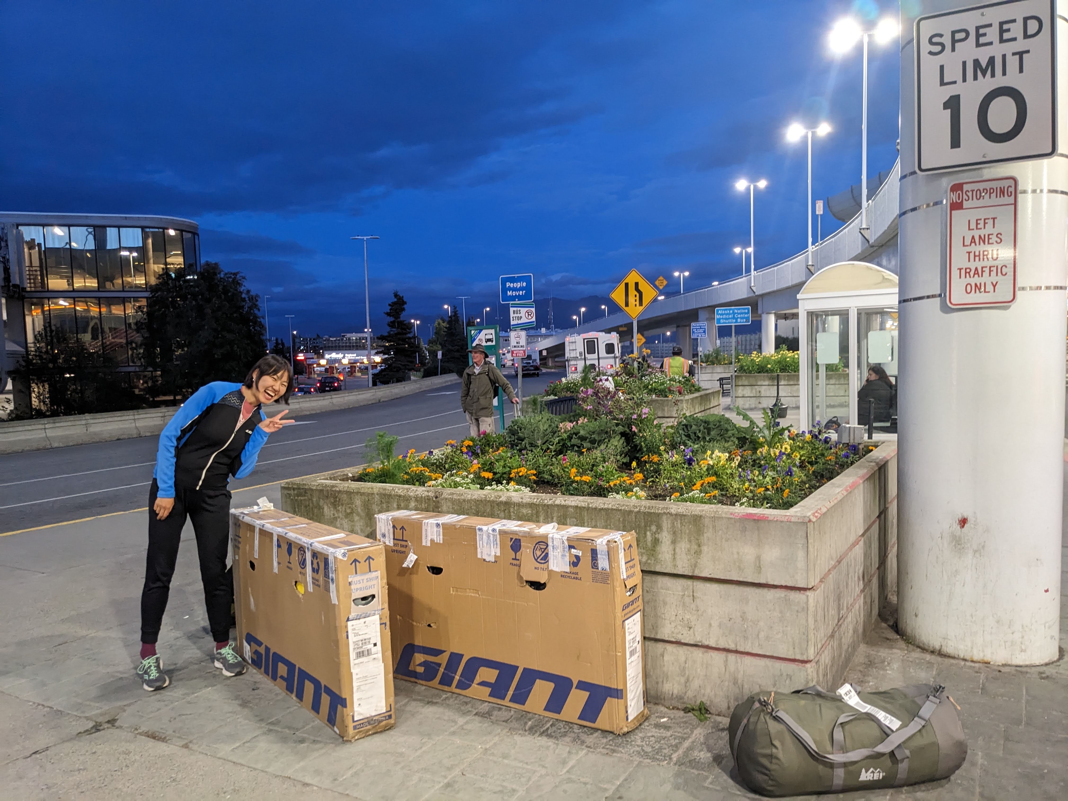 lucy making a peace sign standing over two cardboard bike boxes outside of the anchorage airport terminal