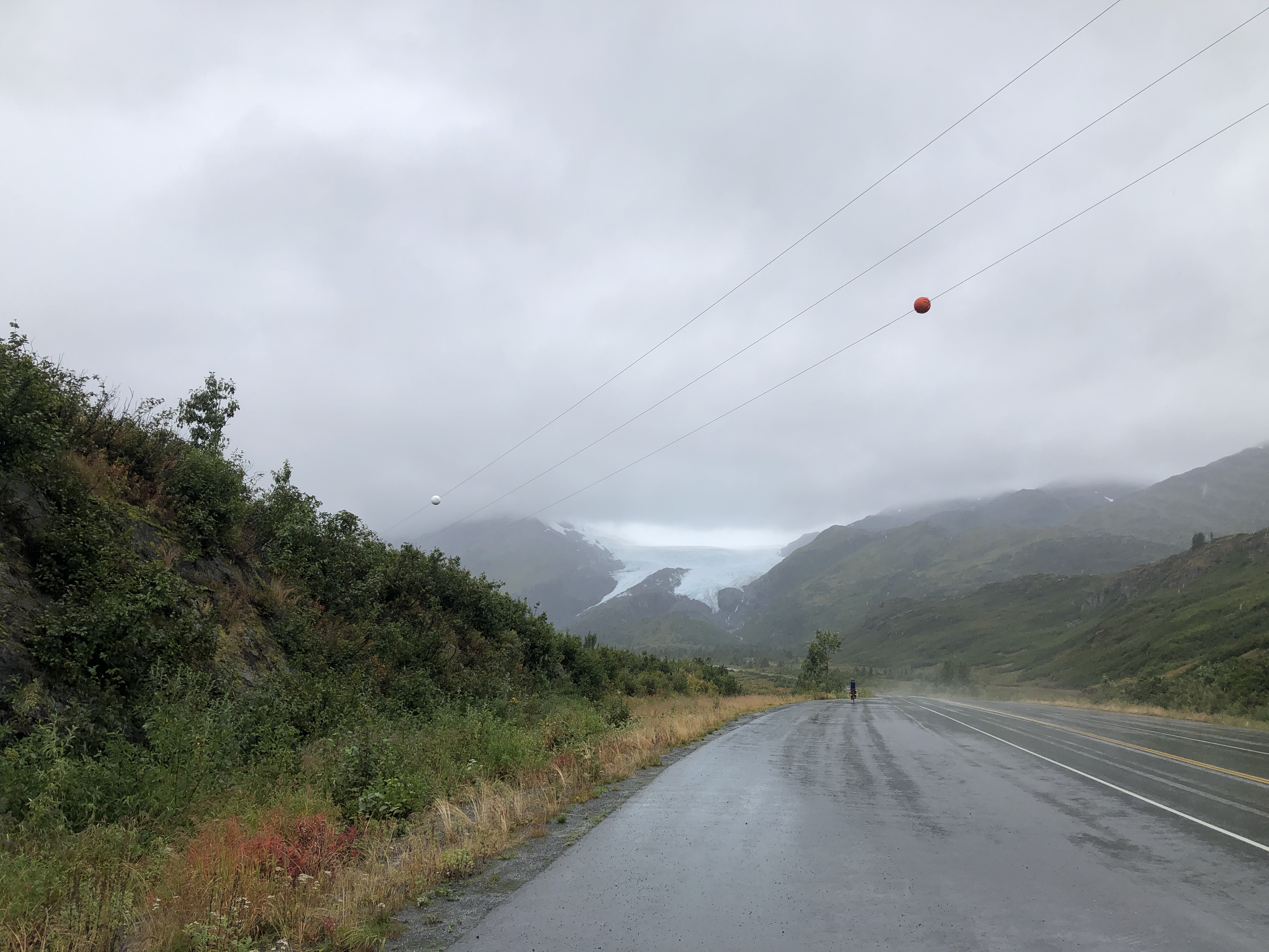 wet road with a cyclist in the distance and glaciers in the background